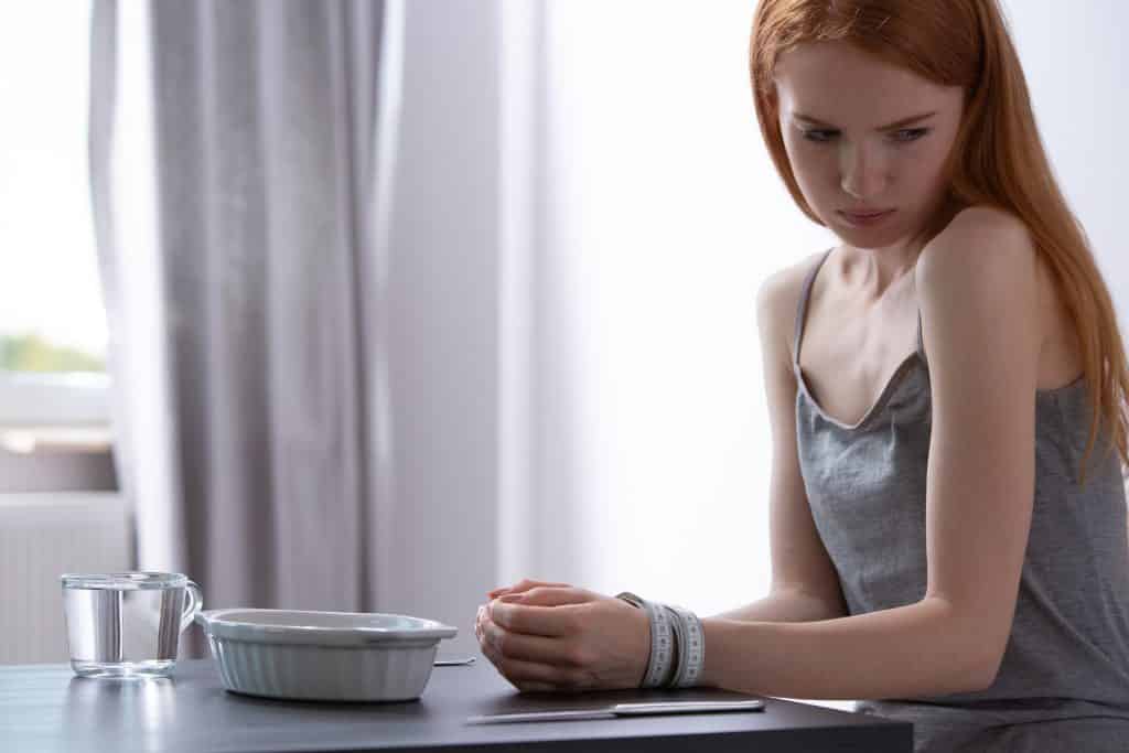 Imagem de uma jovem mulher que sofre de anorexia. Ela veste uma regata cinza. Esta sentada à frente de uma mesa e suas mãos estão amarradas com uma fita métrica.
