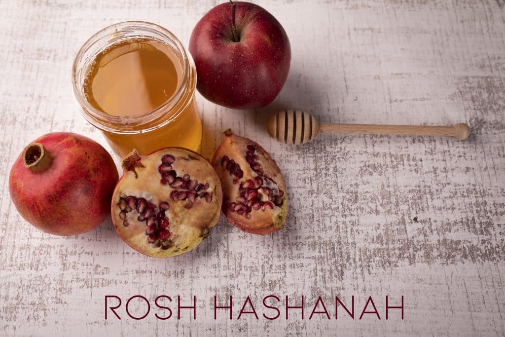 Imagem de frutas como romã e maçã, um copo de mel e o seu dosador, elementos que compõem a celebração do ano novo judaico. À frente dos ingredientes está escrito a palavra: Rosh HaShaná 
