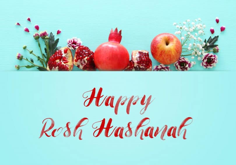 Imagem de várias frutas como romã e maçã decoradas com ramos de flores para compor os rituais para celebrar o ano novo judaíco. À frente dos elementos está escrito em inglês a frase: Happy Rosh HaShaná.