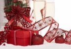 Imagem de uma mesa decorada com vários itens para celebrar o natal e o ano novo. Entre eles, descatam-se: uma caixa de presentes na cor vermelha, laçcos de fita com flores vermelhras, bolas de natal e duas taças de champanhe.