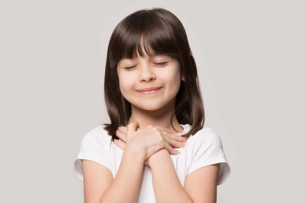 Menina com as mãos no peito representando a gratidão com uma blusa branca no fundo cinza