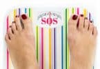 Imagem de uma balança de peso de banheiro colorida e no visor dos números está escrito a palavra SOS em cor rosa. Sobre a balança os pés de uma mulher com as unhas pintadas de vermelho escuro.