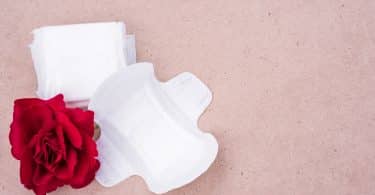 Imagem de dois absorventes femininos sem uso. Um aberto e o outro fechado. Ao lado deles uma rosa vermelha.