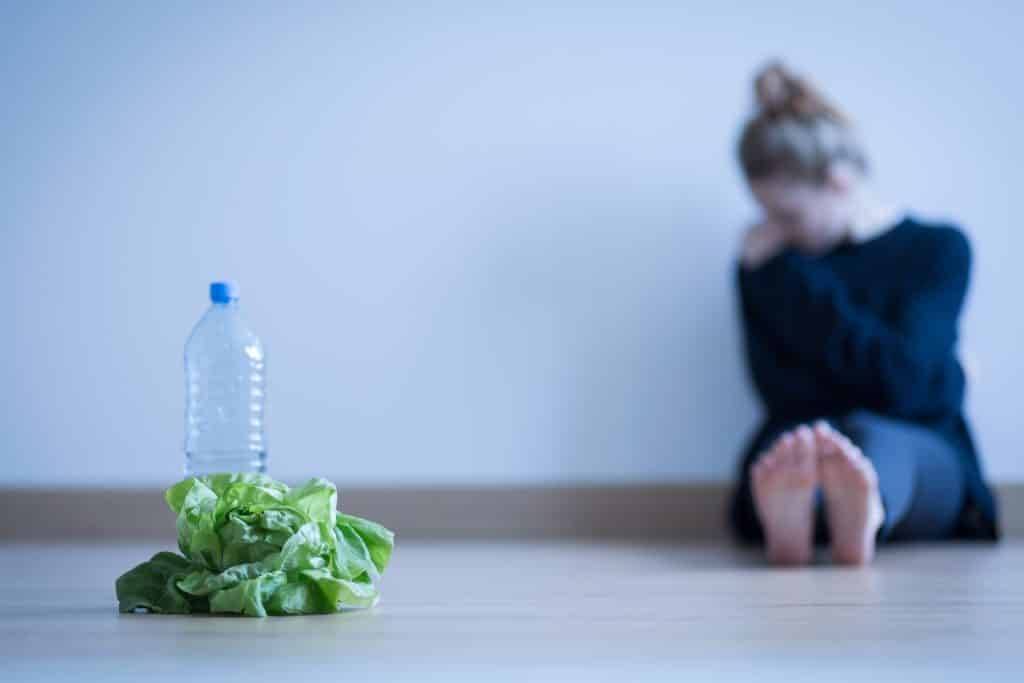 Imagem de uma sala vazia. Ao fundo uma mulher vestida com roupa preta sentada ao chão. Ela sofre de anorexia. À sua frente, um maço de alface e uma garrafa plástica com água.
