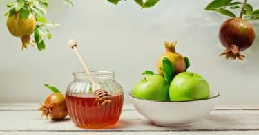 Imagem de uma mesa de madeira na cor branca e sobre ela uma tigela com maças verdes e romã, um pote com mel e uma romã ao lado do pote. Esses são alguns alimentos para celebrar o ano novo judaíco.