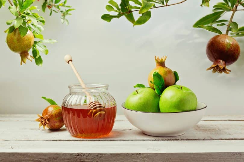 Imagem de uma mesa de madeira na cor branca e sobre ela uma tigela com maças verdes e romã, um pote com mel e uma romã ao lado do pote. Esses são alguns alimentos para celebrar o ano novo judaíco.