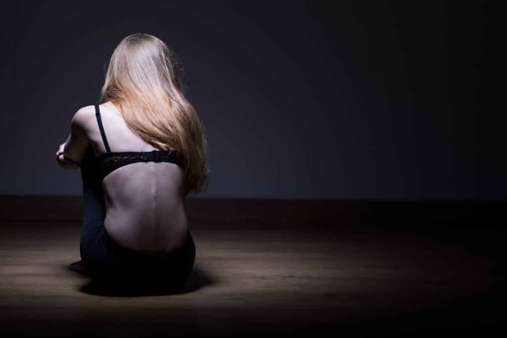 Imagem de uma jovem mulher sentada de costas, olhando para uma parede. Ela sofre de anorexia. Usa uma calça preta e um sutiã também na cor preta.
