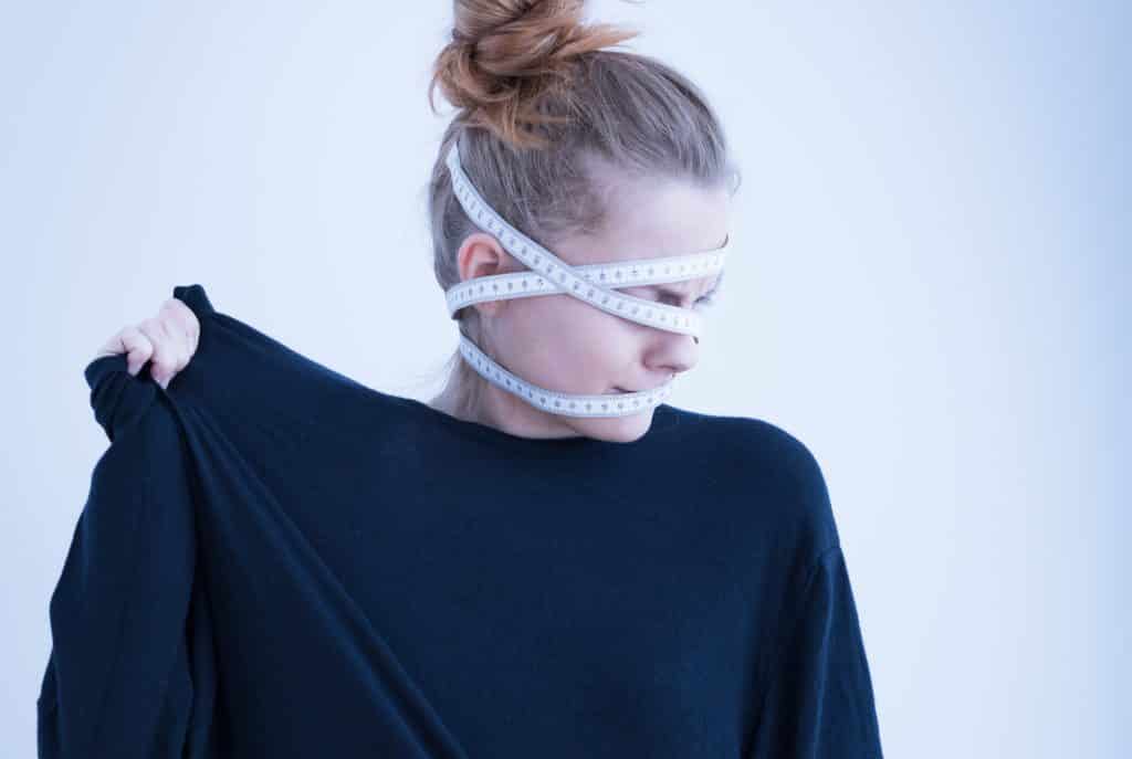 Imagem de uma mulher usando uma blusa de manga longa preta. Ela sofre de anorexia e está com uma fita métrica enrolada em sua cabeça.