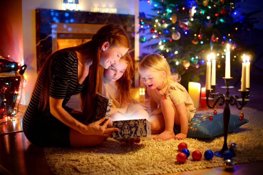Uma mulher e duas crianças pequenas abrindo um presente de Natal perto de uma árvore de Natal