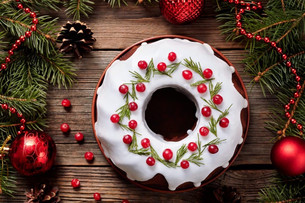 Imagem de um lindo e delicioso bolo de natal, coberto com glacê, decorado com cerejas e ramos verdes. O bolo está disposto sobre uma mesa de madeira, decorada com bolas vermelhas de natal.