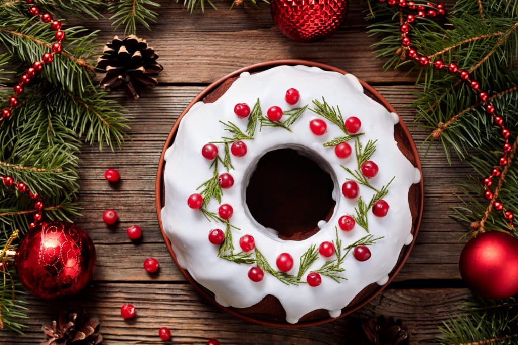 Imagem de um lindo e delicioso bolo de natal, coberto com glacê, decorado com cerejas e ramos verdes. O bolo está disposto sobre uma mesa de madeira, decorada com bolas vermelhas de natal.
