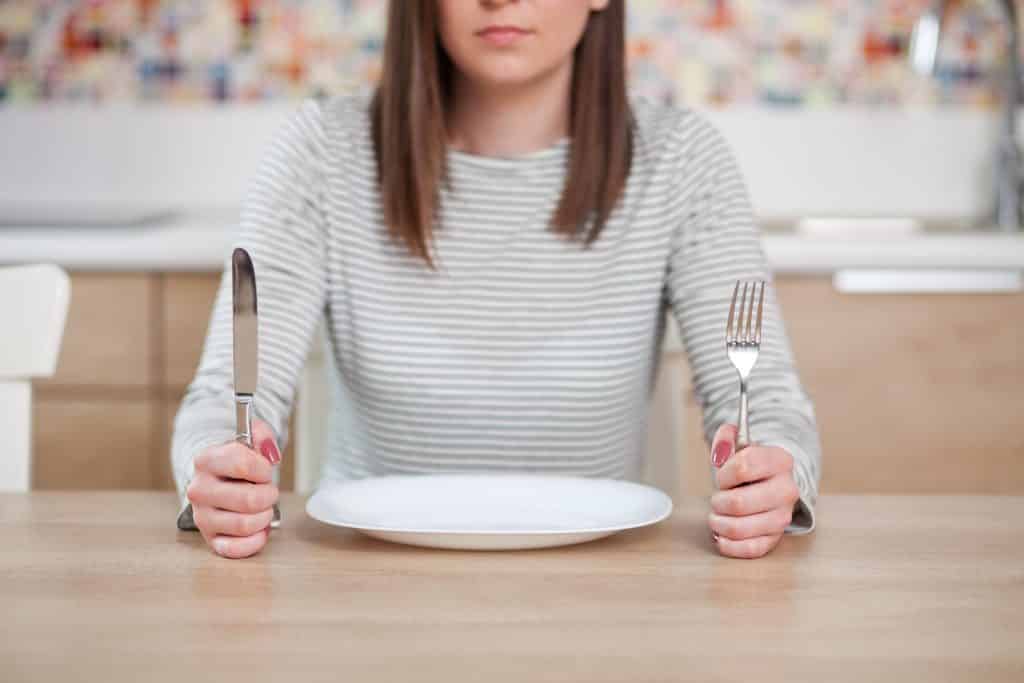 Imagem de uma moça usando uma blusa com listras branca e cinza de manga longa sentada à frente de um prato vazio. Em suas mãos ela segura um garfo e uma faca.
