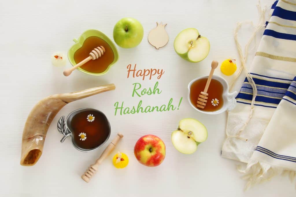 Imagem de vários elementos como maçãs, mel, dosador, que compõem a celebração do ano novo judaíco. No centro desses elementos está escrito a palavra: Happy Rosh HaShaná em inglês.