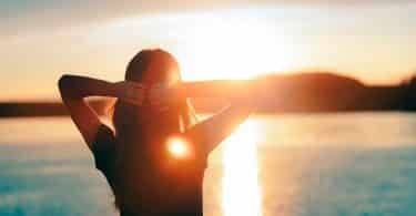 Mulher com os braços erguidos olhando para o pôr do sol refletido na água.