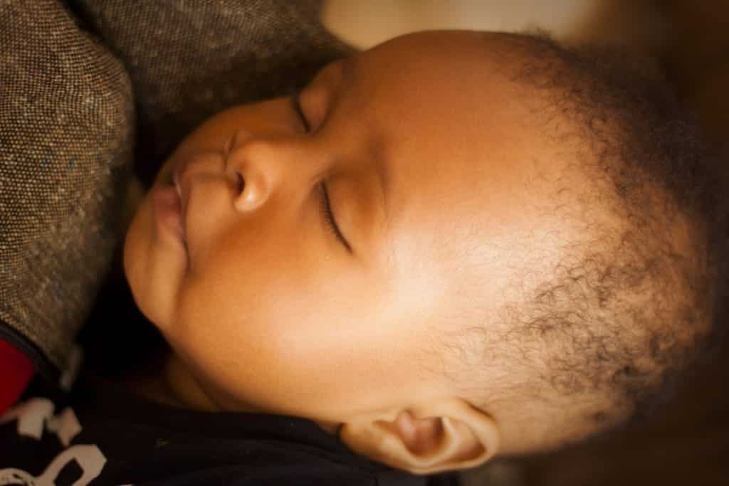 Imagem de um lindo recém nascido com poucos cabelos encaracolados. Ele está dormindo no colo de sua mãe.
