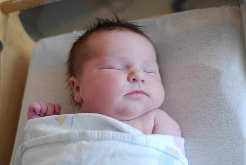 Imagem de um lindo recém nascido envolto de uma matinha. Ele está dormindo em um colchão forrado com um lençol bege bem clarinho.
