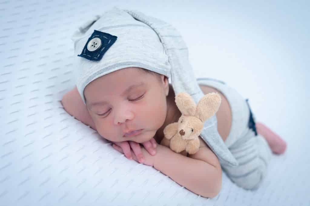 Imagem de um lindo bebê recém nascido dormindo. Ele está deitado de bruços sobre uma cama com lençol branco. Ele usa uma touca cinza moderna e ao lado dele um coelhinho de pelúcia complementa a imagem.