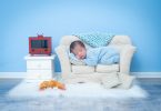 Imagem de um quarto infantil decorado com uma poltrona brnaca, um tapete de pelo, um criado de duas gavetas com uma TV de pequena polegada sobre ele. No sofá temos um recém nascido dormindo bem gostoso.