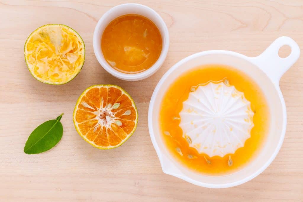 Imagem de um suco detox de laranja em uma jarra. Ele está sendo coado. Ao lado dele uma laranja cortada ao meio e um copo com o suco.

