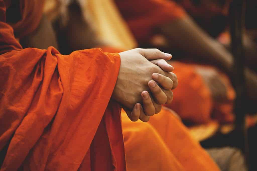 Imagem de um monge sentado e meditando. Ele veste um longo vestido alaranjado e está com as suas mãos entrelaçadas.