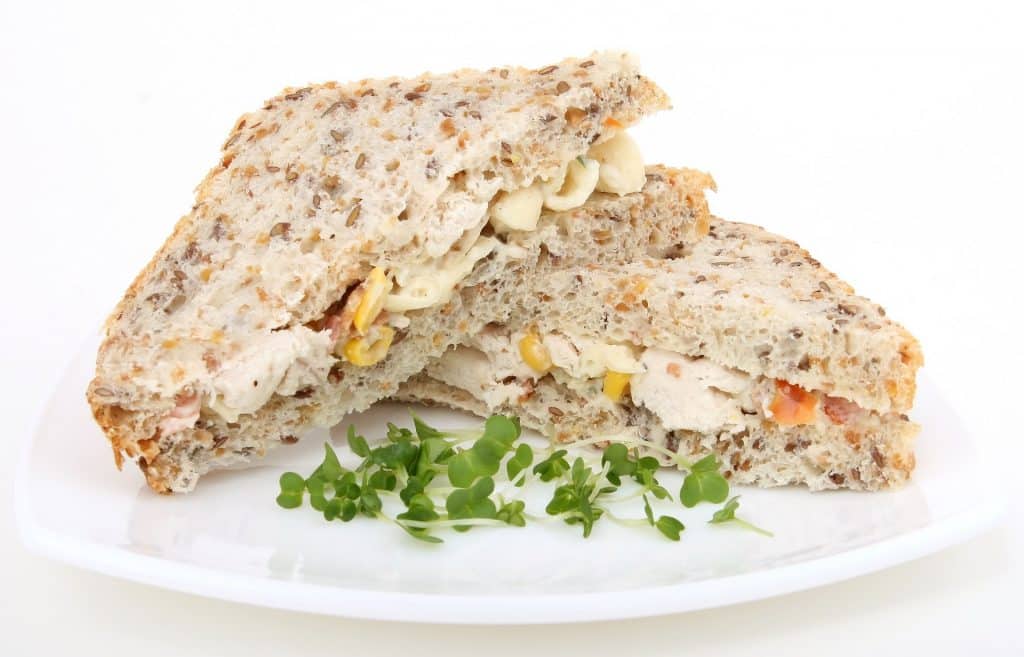 Imagem de um delicioso lanche de peru, feito com as sobras da ceia de natal. O lanche é feito em pão de forma e está dividido ao meio, disposto em um prato, decorado com ramos de orégano frecos.