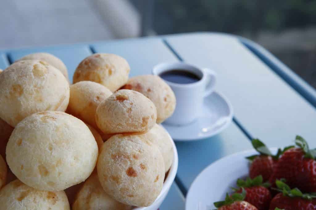 Imagem de um prato com pão de queijo feito com batata-doce. Os pães já estão assados. Ao lado do prato uma xícara de café.
