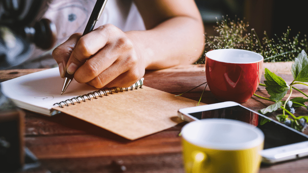 Mulher trabalhando e escrevendo em caderno com canecas sobre a mesa