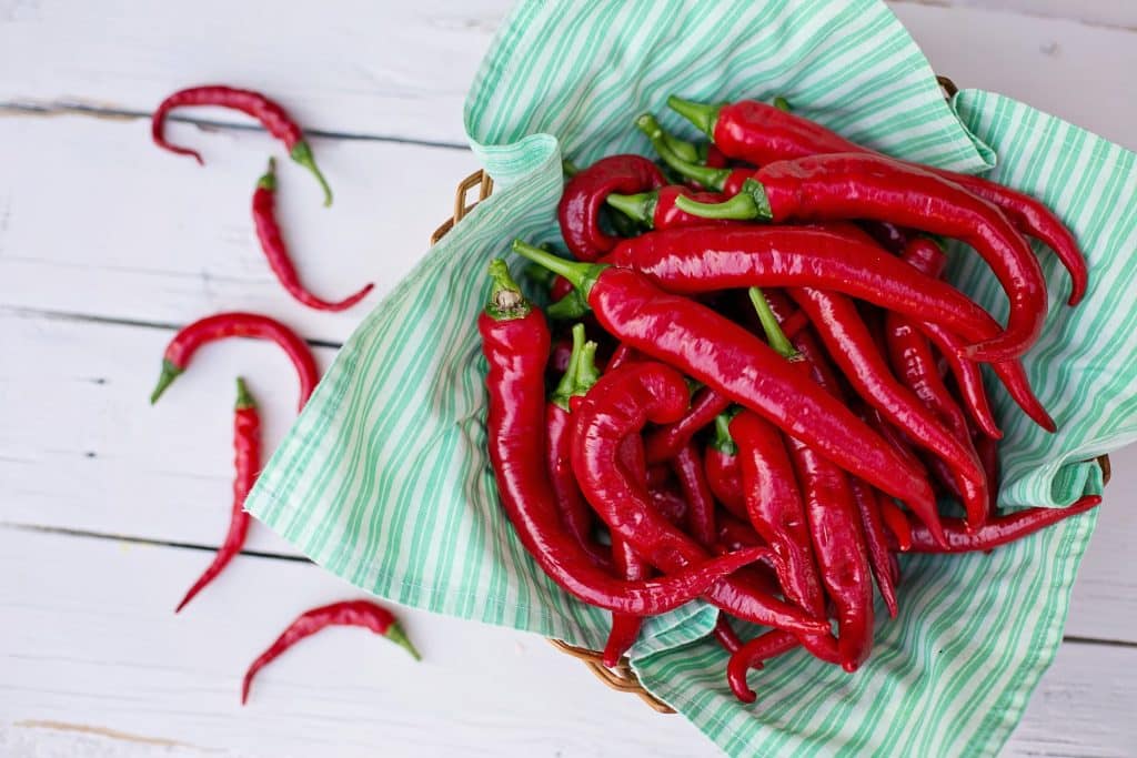 Imagem de várias pimentas caienas bem vermelhas dispostas sobre um cesto de vime coberto com uma toalha branca com listras verdes. Essa pimenta é indicada para dor na garganta.