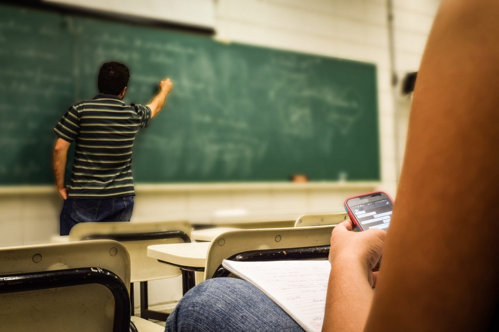 Imagem de uma sala de aula. Ao fundo um quadro negro e o professor escrevendo nele. Ao fundo a imagem de uma pessoa mexendo no celular e não prestando atenção no ensinamento do professor.