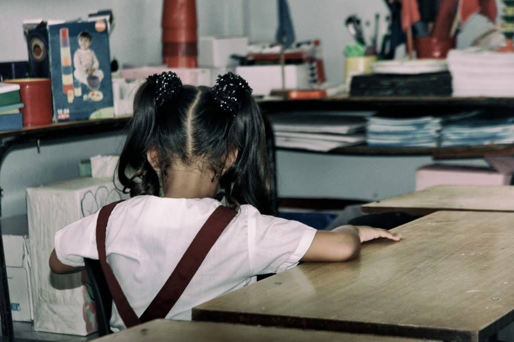 Imagem de uma criança em uma sala de aula. Ela está sentada de costas. Usa o uniforme escolar com camisa branca e jardineira na cor vinho. 