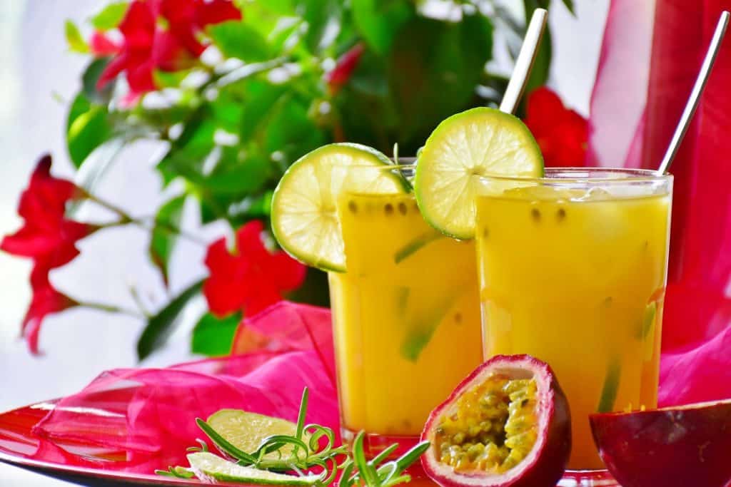 Imagem de dois copos de suco detox feitos com frutas amarelas. Os copos estão decorados com fatias de limão e estao sobre uma mesa decorada com um tecido de voal na cor rosa.