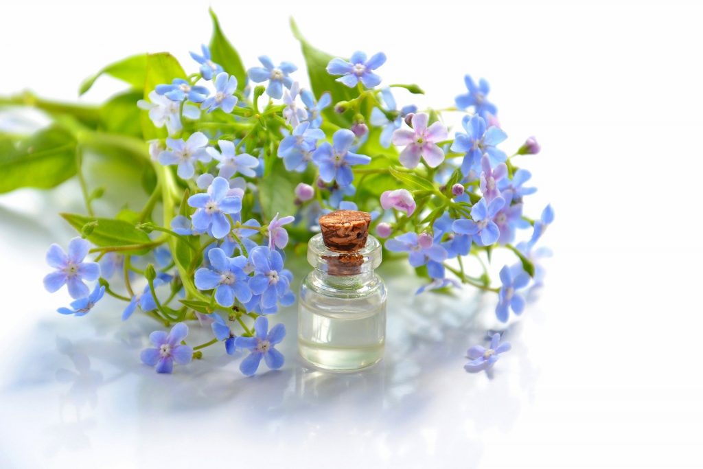 Imagem de um pequeno frasco contendo óleos essenciais. Ao lado um ramo de flores pequenas na cor azul.