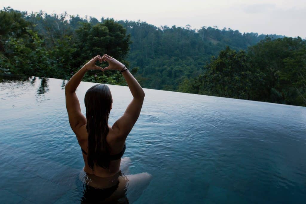 Imagem de uma mulher dentro de uma piscina de borda infinita. Ela está olhando para uma montanha. Está com os braços erguidos e com as mãos juntas, está formando a imagem de um coração.
