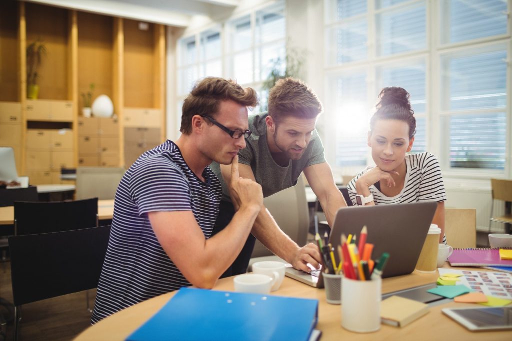 Imagem de três pessoas, dois homens e uma mulher em uma sala com vários armários, mesas, cadeiras. Eles estão reunidos em uma das mesas olhando para o notebook. 
