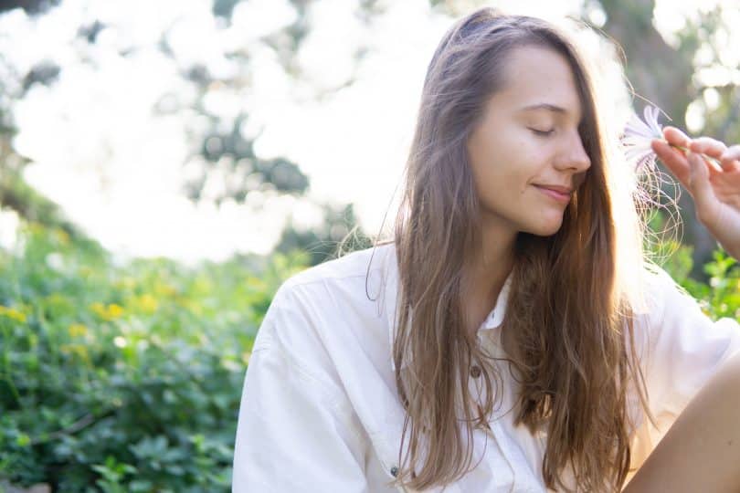 Mulher sentada em um jardim de olhos fechados segurando uma flor