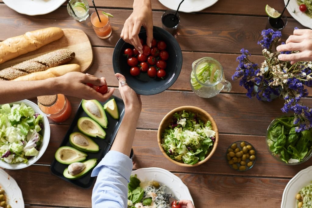 Mesa repleta de alimentos enquanto pessoas mexem neles.