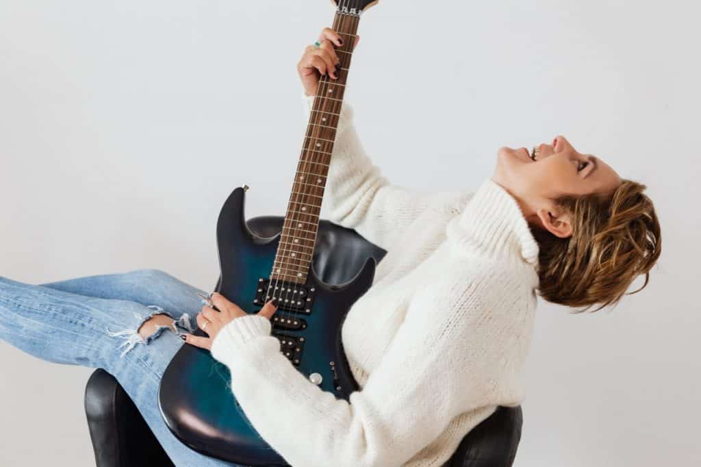 Mulher branca e loira de cabelos curtos, sentada numa poltrona preta, segurando guitarra enquanto olha para cima.