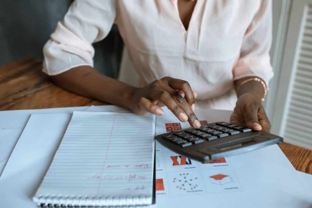 Mulher negra mexendo na calculadora com vários papéis sobre finanças em cima da mesa 