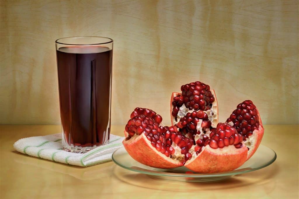 Imagem de um copo de suco detox feito com frutas vermelhas. Ao lado do copo, um pires contendo uma romã cortado em quatro partes iguais