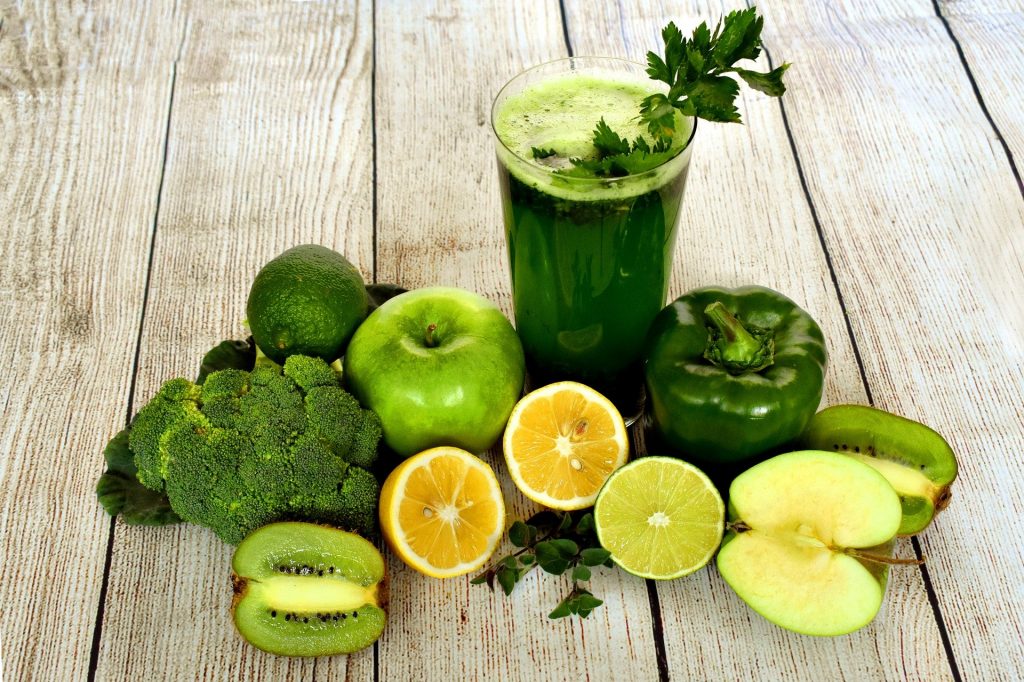 Imagem de um copo contendo suco detox verde. O copo é decorado com salsinha e ao lado vários legumes, frutas e verduras verde, utilizados para o preparo do mesmo.