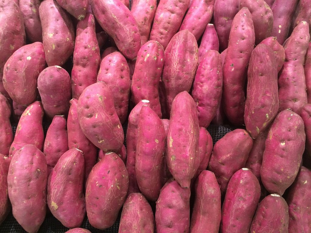 Imagem de uma bancada de feira contendo muitas batatas doces da cor roxa.