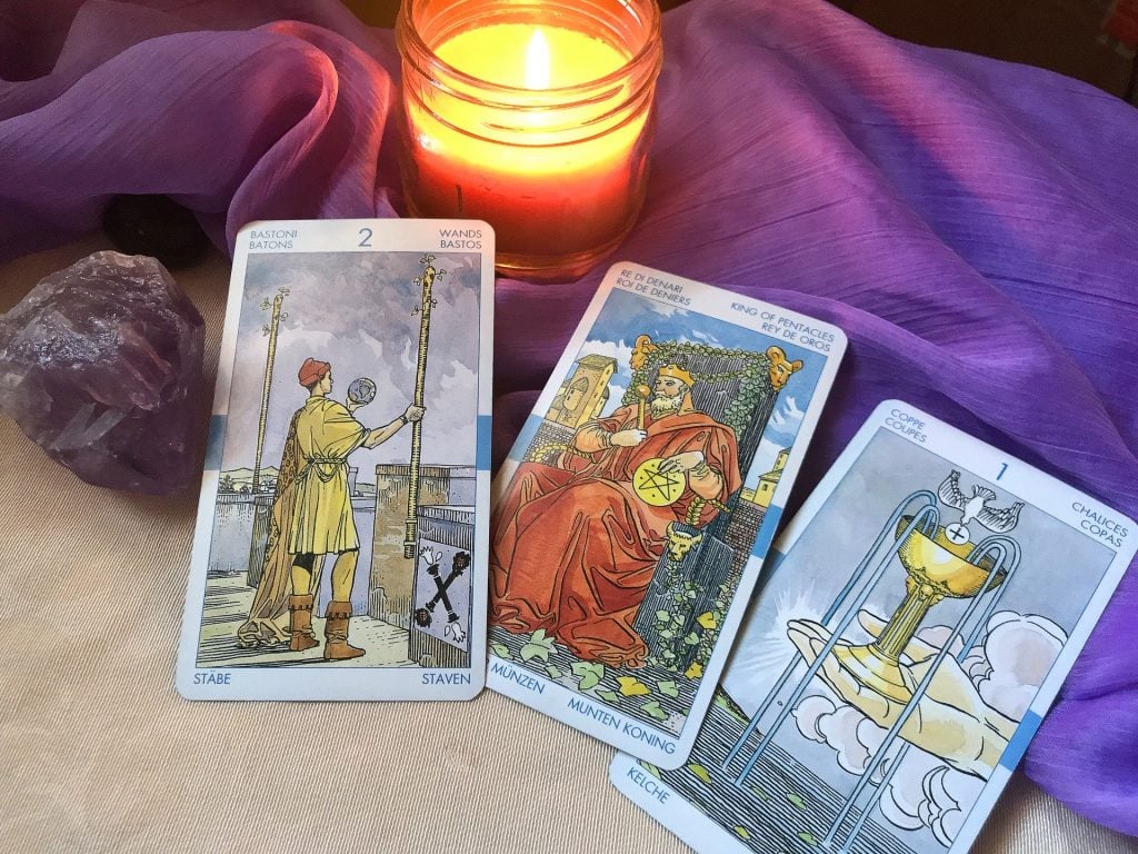 Imagem de uma mesa forrada com um tecido roxo e sobre ela três cartas do tarot. Ao lado uma pedra lilás e um pote com uma vela acesa.
