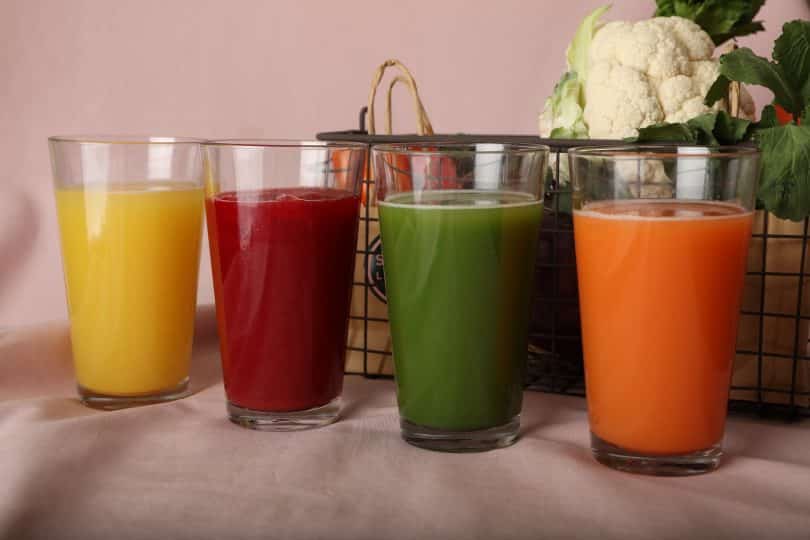 Imagem de quatro copos contendo vários tipos de sucos detox. Ao lado dele um cesto com ingredientes como: frutas e legumes, utilizados para o preparo dos mesmos.