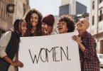 Mulheres com expressões sorridentes segurando cartaz com a palavra "women".