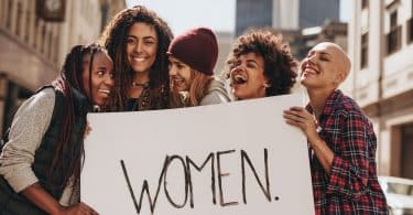 Mulheres com expressões sorridentes segurando cartaz com a palavra "women".