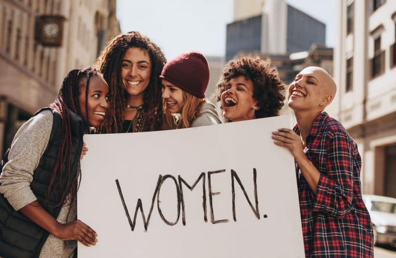 Mulheres com expressões sorridentes segurando cartaz com a palavra "women".