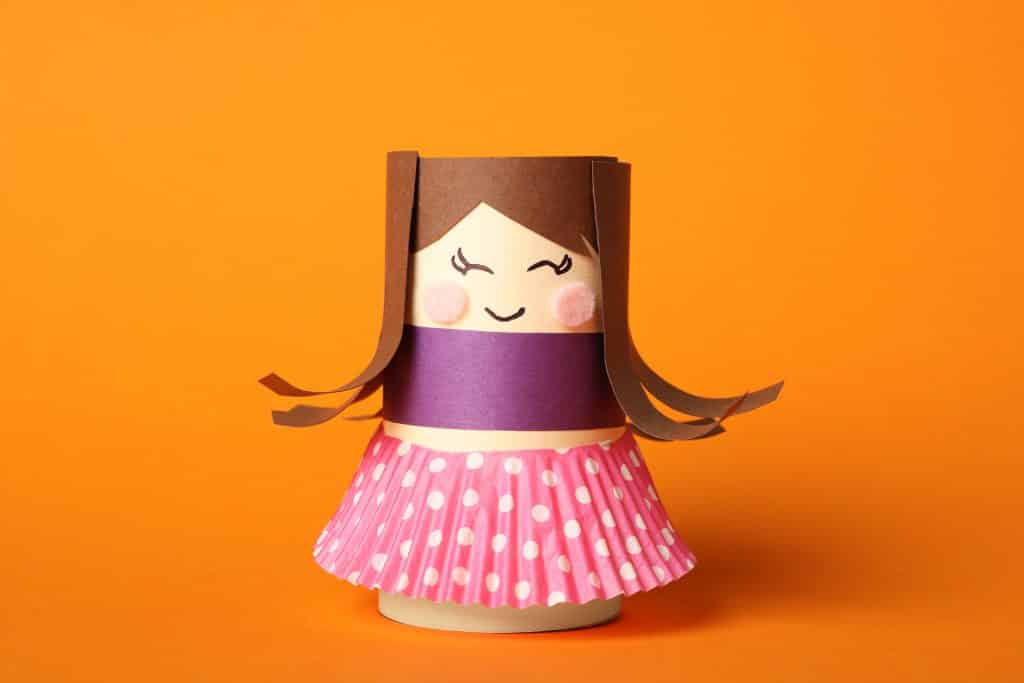 Imagem de um brinquedo reciclado feito de rolo de papel higiênico. É uma bonequinha bem colorida usando uma sainha rosa de bolinha branca.