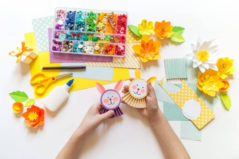 Imagem de uma mesa com vários itens para a fabricação de brinquedos recicláveis. A mão de uma criança segura dois bonecos já prontos.