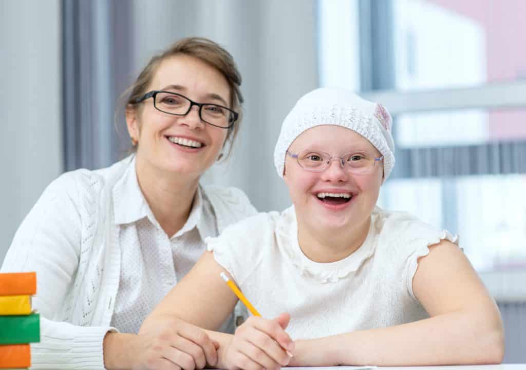 Imagem de uma criança portadora da síndrome de down em uma sala de aula. Ela está ao lado da sua professora. Ela segura um lápis de cor amarela. Ambas usam óculos de grau e roupas brancas e estão sorrindo.