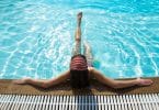 Imagem de uma piscina com uma água azul e limpa. Dentro dela uma mulher encostada na beirada relaxando as suas pernas.